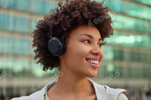 Preview: Headshot Of Positive Woman With Curly Hair Smiles Happily Listens Audio Track Via Headphones