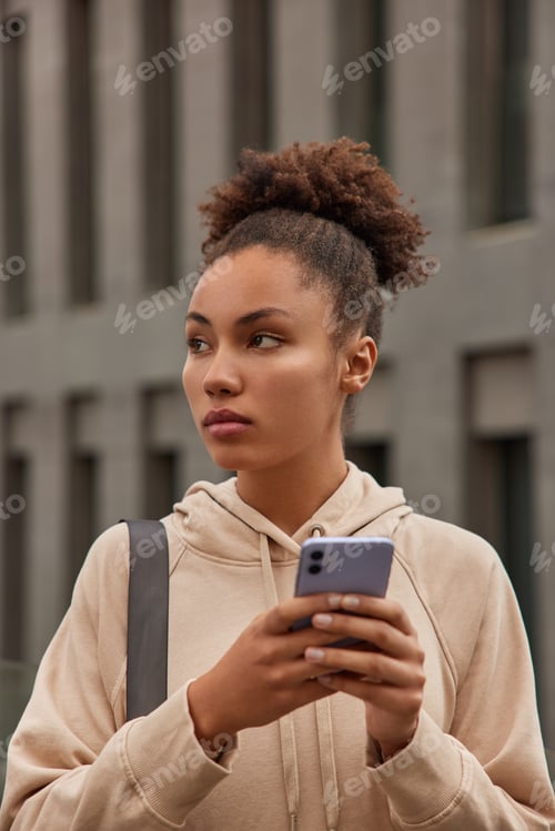 Preview: Photo Of Curly Haired Afro American Woman Dressed In Hoodie Holds Smartphone Checks Text Message