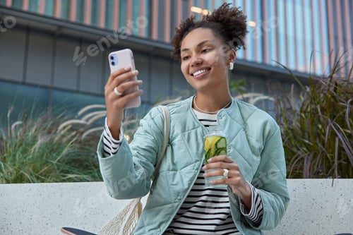 Preview: Outdoor Shot Of Happy Teenage Girl With Two Curly Buns Takes Selfie Via Smartphone Drinks Detox