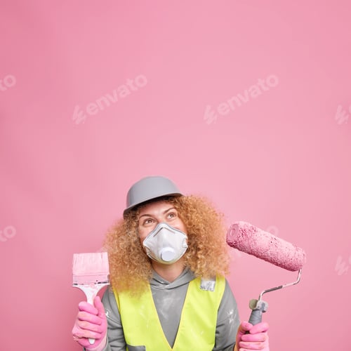 Preview: Pleased Curly Female Builder Or Laborer Wears Helmet And Protective Respirator Unform Holds Paint