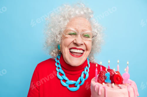 Preview: Positive Aged Woman With Grey Curly Hair Smiles Broadly Has White Perfect Teeth Holds Cake