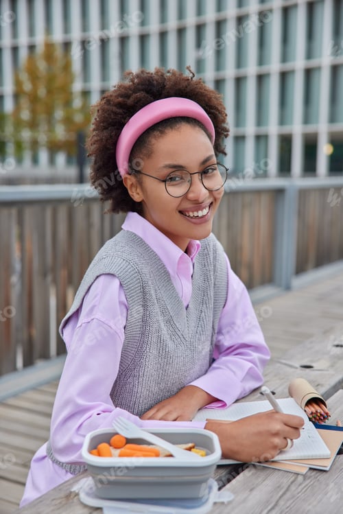 Preview: Positive Creative Woman Makes Notes Poses At Wooden Table With Lunch Box Outdoors In City Has Very
