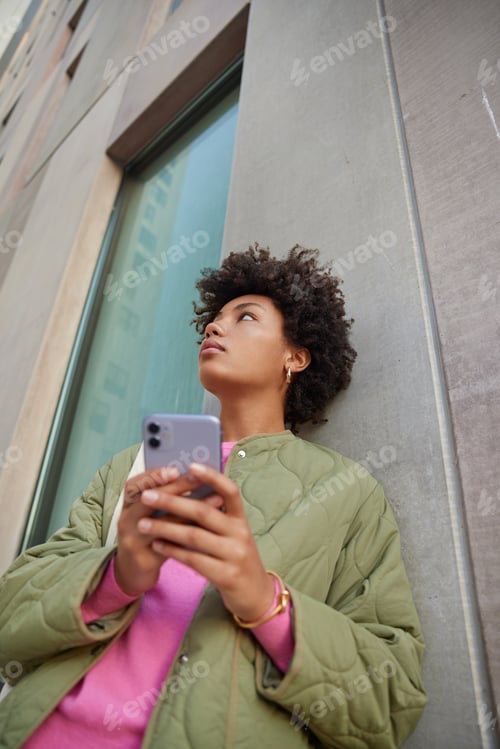 Preview: View From Below Of Thoughtful Curly Haired Woman Uses Phone Chatting With Fried Holds Mobile Phone