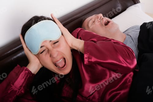 Preview: Portrait Of Irritated Woman Blocking Ears With Hands While Man Snoring On Bed. Indoors