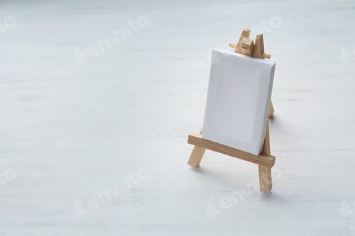 Preview: Close Up View Of Blank Canvas Miniature With Easel On White Wooden Background. Art Minimal Concept.