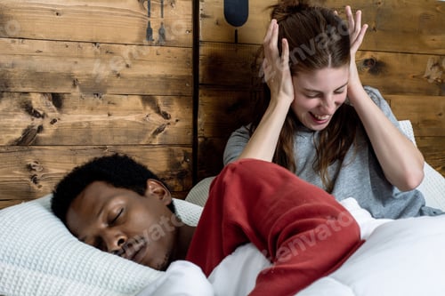 Preview: Portrait Of Irritated Woman Blocking Ears With Hands While Man Snoring On Bed. Multiracial Couple