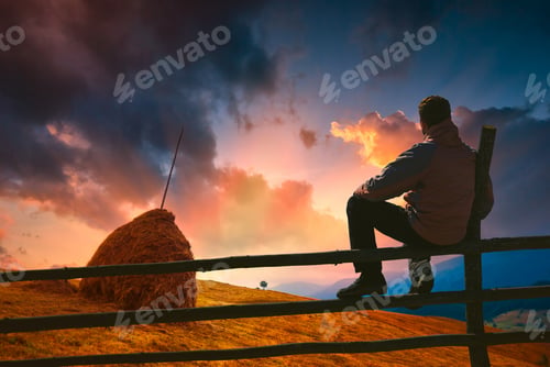 Preview: Man Sitting On A Wooden Fence And Enjoy Wonderful Bright Sunset With Majestic Colorful Clouds In A