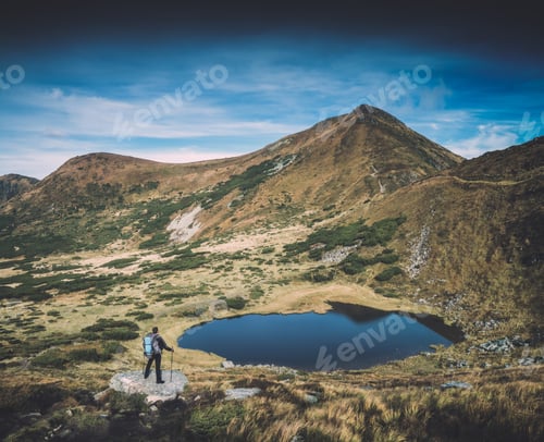 Preview: Hiker With Backpack And Trekking Poles Standing On A Big Stone Near The Mountain Lake In A