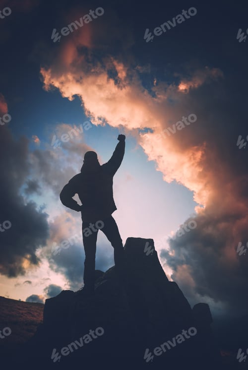 Preview: Man Standing On A Cliffs Edge With Raised Hand Against Colorful Clouds In A Light Of Sunset