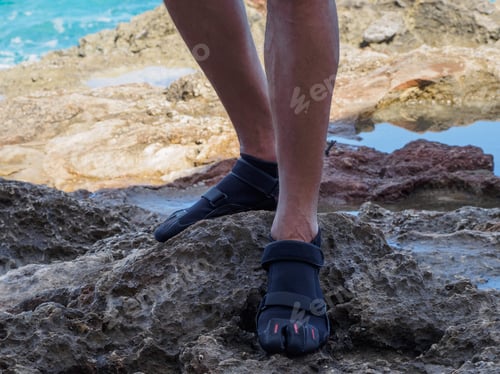 Preview: Crop A Man In Reef Shoes Is Standing On A Reef. Sharp Stones. Near The Ocean.
