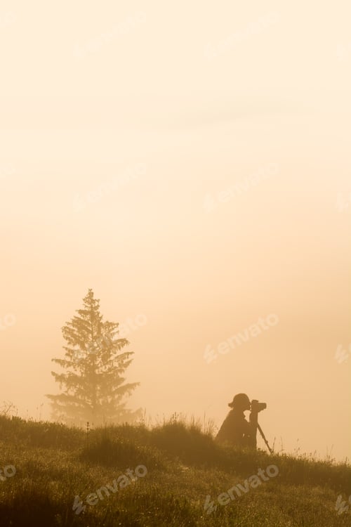 Preview: Silhouette Of A Woman Photographer Sitting On A Grass In A Misty Valley. Early Morning Time.