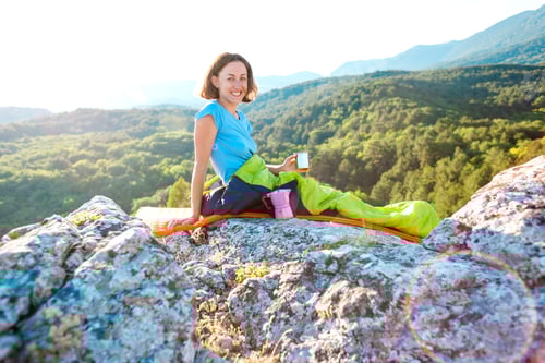Preview: A Woman Is Drinking Coffee While Sitting On Top Of A Mountain. A Girl In A Sleeping Bag Drinks A