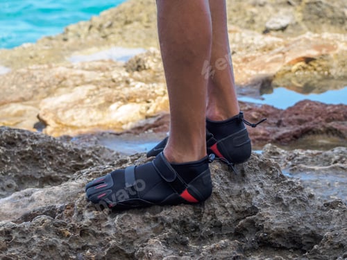 Preview: Crop A Man In Reef Shoes Is Standing On A Reef. Sharp Stones. Near The Ocean.
