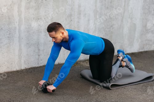 Preview: Muscular Bodybuilder Guy In A Blue T-Shirt Pumping Abs With Roller On Gray Carimate. Fitness And