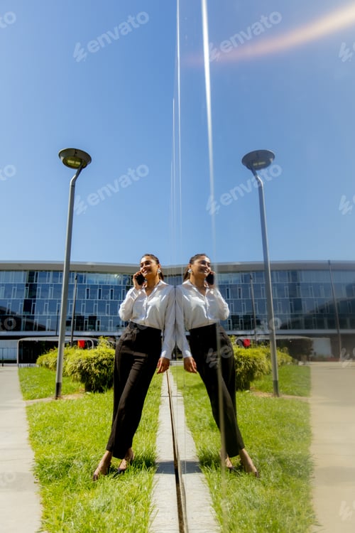 Preview: Businesswoman talking on phone near building reflecting in glass