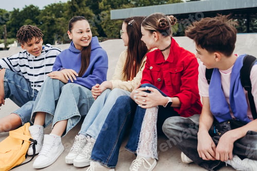 Preview: Group of happy teenage friends sitting together at skatepark and talking outdoors