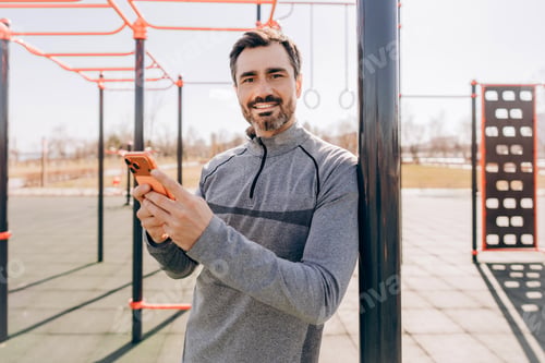 Preview: Smiling sporty man resting after workout and using smartphone at outdoor fitness playground