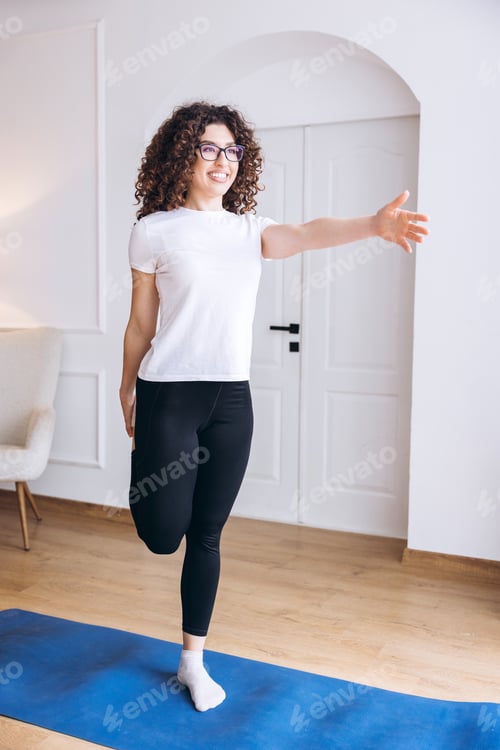 Preview: Happy, smiling young woman stretching leg quad on blue yoga mat at home