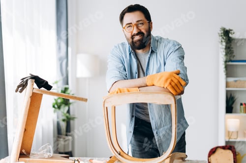 Preview: Smiling man wearing gloves repairing old wooden chair looking at camera