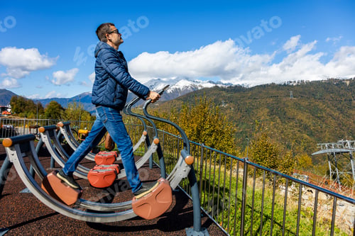 Preview: Man uses outdoor exercise machine on mountain with snow-capped peaks