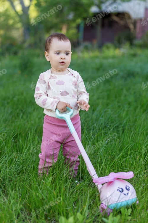 Preview: Toddler girl walking in green grass with push toy outdoors, active child lifestyle