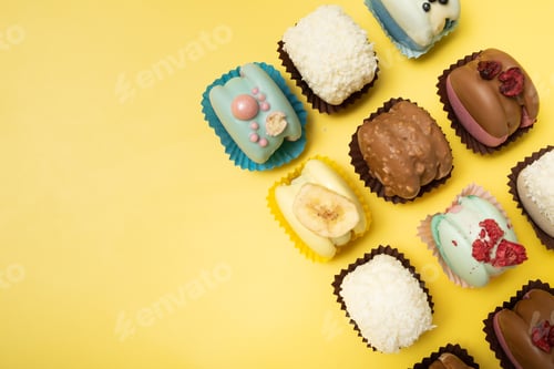Preview: Colorful small cakes arranged on a yellow background in different shapes and flavors with decorative