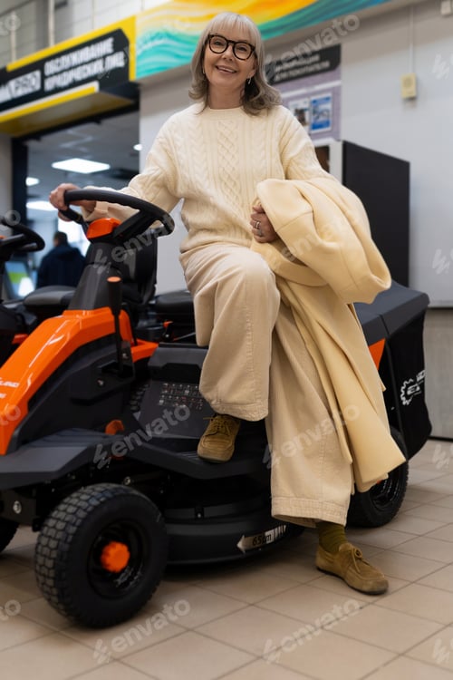Preview: Senior woman in glasses smiling while sitting on an orange riding lawn mower in a retail store