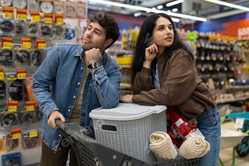 Preview: Couple pushing shopping cart and thinking about options in a brightly lit hardware store aisle