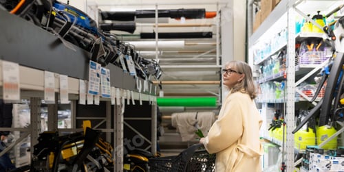 Preview: Senior woman shopping for products in a hardware store aisle with a cart
