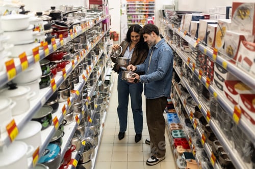 Preview: Young couple comparing cooking pots in a store aisle, buying household items together