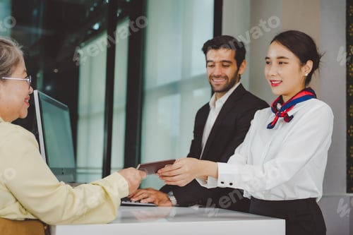Preview: Airline ground staff handing passport to senior woman at airport desk with friendly business travele