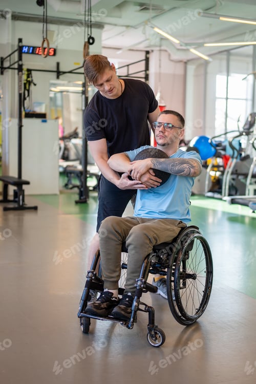 Preview: Personal coach supporting disabled man in wheelchair during power workout with medicine ball in gym.