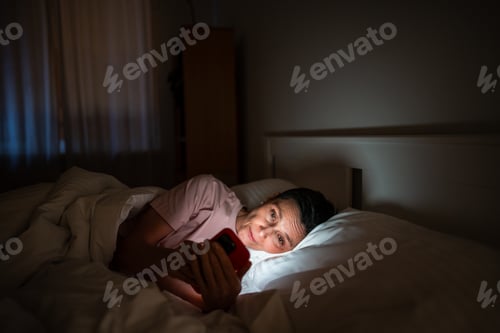 Preview: Sleepy senior woman phone addict scrolling social media online on smartphone late at night.