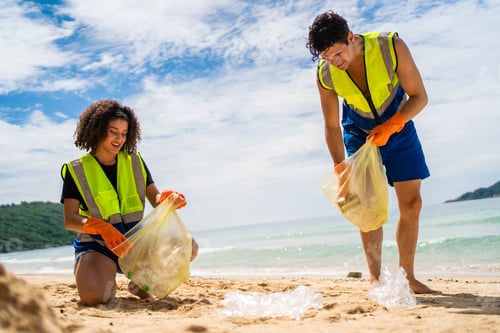 Preview: Two volunteers collecting plastic bottles and trash on a sandy beach.