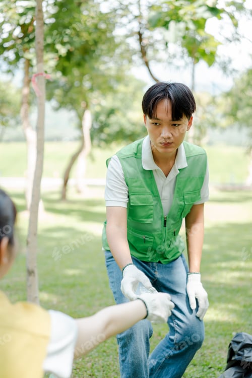 Preview: Young asian man in a green vest collecting plastic waste into a black bag outdoors, accepting more t
