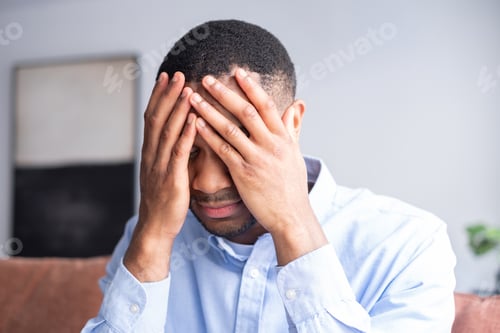 Preview: Stressed young adult african american man suffering from a headache, covering his face with hands, f