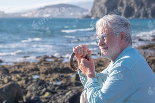 Preview: Portrait of pensive bearded senior man resting at the beach sitting with hands on a walking cane. Ha