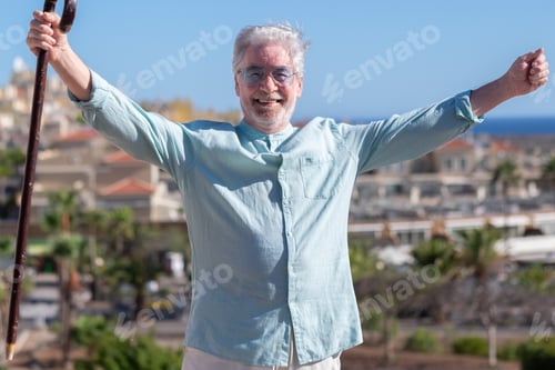 Preview: Cheerful white-haired senior man outdoors in a sunny day holding a walking cane looking at camera wi