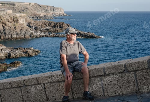 Preview: Senior man sitting on a stone wall by the ocean, contemplating the vast sea view, enjoying a peacefu