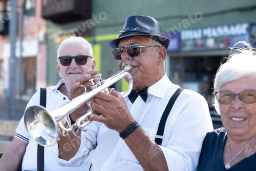 Preview: Senior man playing a trumpet while wearing a fedora, bowtie, and suspenders, entertaining an appreci