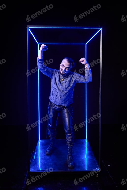 Preview: Young man with skeleton face paint posing confidently inside neon blue light box in dark studio