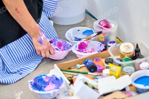 Preview: Artist hand mixing bright acrylic paints on floor surrounded by brushes and containers