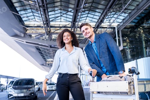 Preview: Smiling couple with luggage cart at modern airport terminal entrance