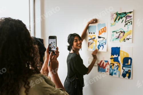 Preview: Friend taking a photo with phone camera of her artist friend by a mood board on a white wall