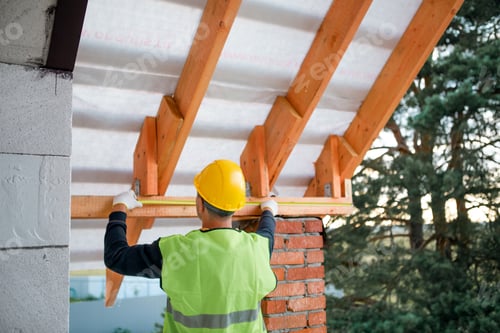 Preview: Builder in safety helmet aligns rafter ends, measuring timber lag placement for roof structure, focu