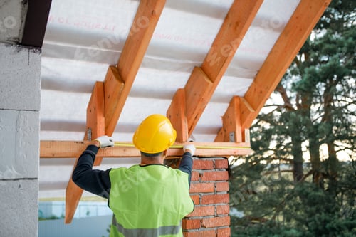 Preview: Male constructor secures ridge beam at roof peak, timber lags visible, advanced roofing in progress,