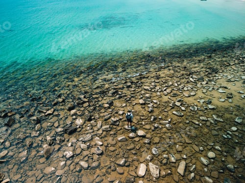 Preview: Top down aerial view of a solo male traveler with a backpack exploring a rocky beach next to clear b