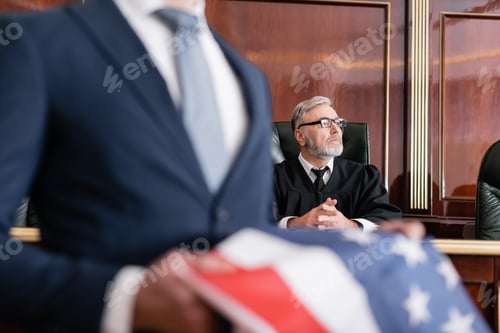 Preview: Senior Judge Looking Away in Courtroom Near Attorney With Usa Flag on Blurred Foreground