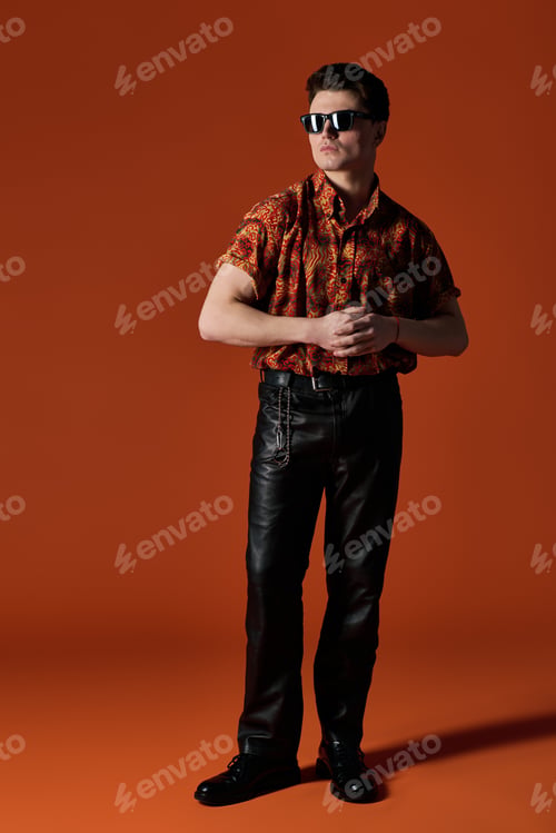 Preview: Handsome young man poses confidently in stylish attire against a vibrant orange backdrop