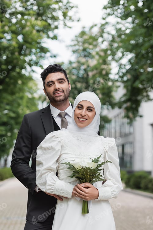Preview: Cheerful Muslim Bride in Wedding Hijab and White Dress Holding Bouquet Near Groom Outside